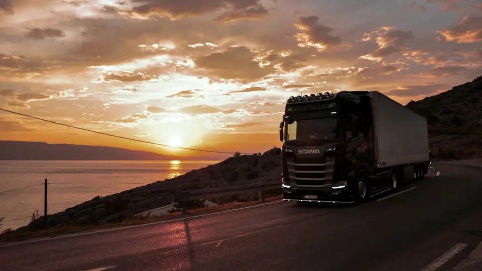A truck with a refrigerated trailer on a coastal road at sunset