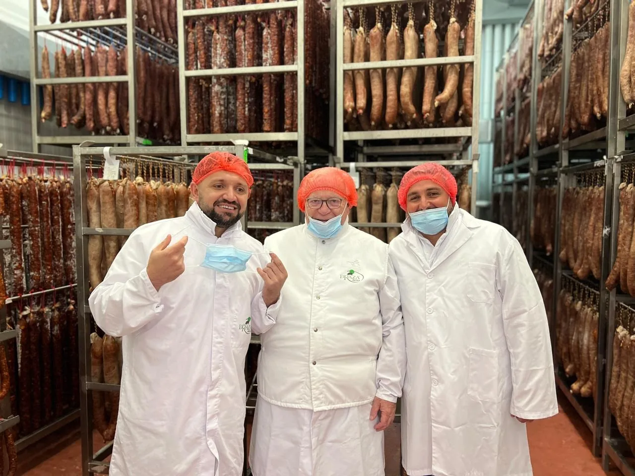 VLC staff in whites, hairnets and masks inside a Spanish curing room, racks of cured meat behind them
